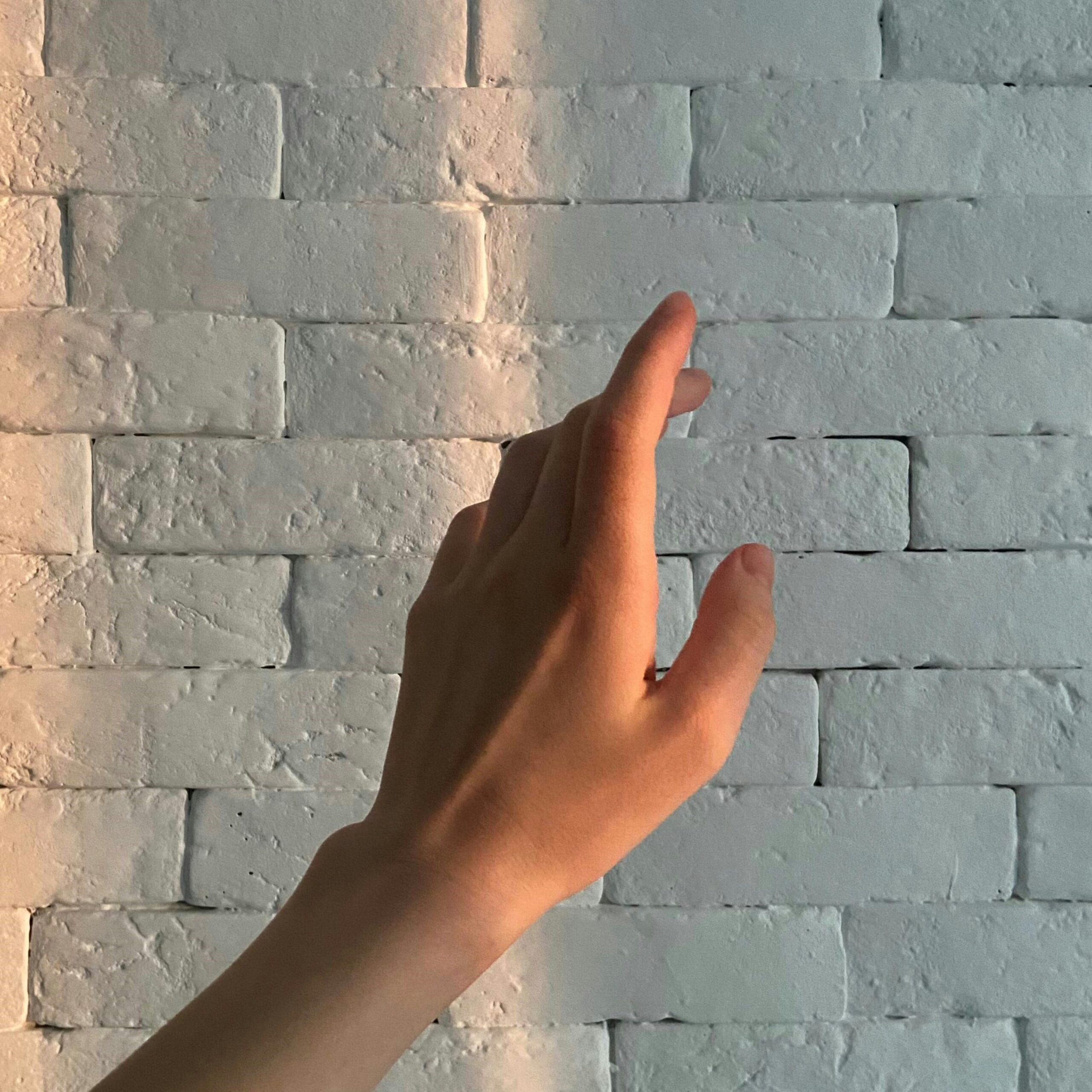 A hand reaching towards a white textured brick wall, captured in warm lighting.